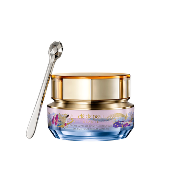 Photo of the 26 AW Holiday Collection limited-edition EYE CONTOUR CREAM SUPREME. The gold-trimmed jar features a blue and pink pastel illustration with a gold shooting star, shown beside a faceted silver spatula.